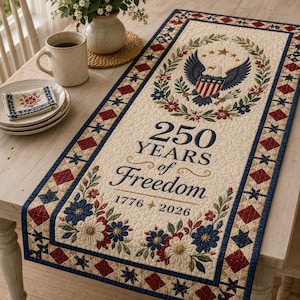 May include: A cream-colored table runner with a patriotic theme. It features a central design of an eagle, stars, and floral elements, with the text "250 Years of Freedom 1776-2026." The border has a repeating pattern of red diamonds and blue stars.