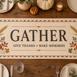 Könnte beinhalten: Ein rechteckiger, gesteppter Tischläufer aus cremefarbenem Stoff mit dem schwarzen Schriftzug GATHER GIVE THANKS MAKE MEMORIES. Das Design zeigt an beiden Enden gestickte orangefarbene Kürbisse, Weizenähren und Herbstblätter.