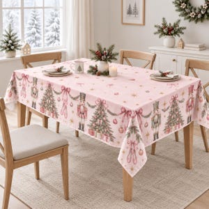 May include: A light pink rectangular tablecloth featuring a repeating Christmas pattern of nutcrackers, decorated pine trees, ballet slippers, and pink bows. The design includes gold star accents and hanging ornaments printed across the fabric surface.
