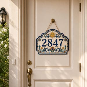 Pode incluir: Uma placa decorativa de cerâmica para número de casa com os dígitos 2847 em azul escuro. A placa tem um fundo creme com um motivo de sol amarelo no topo e padrões florais em azul, verde e laranja. Está pendurada por uma corda castanha.