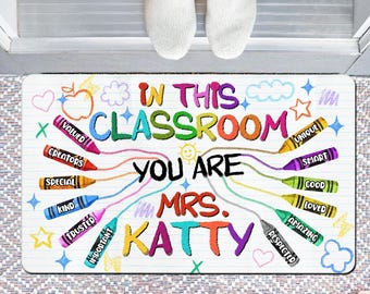 May include: A rectangular doormat with the words "IN THIS CLASSROOM YOU ARE MRS. KATTY" in colourful letters. The design includes crayon illustrations with positive adjectives like "VALUED" and "AMAZING". The mat is on a patterned surface.