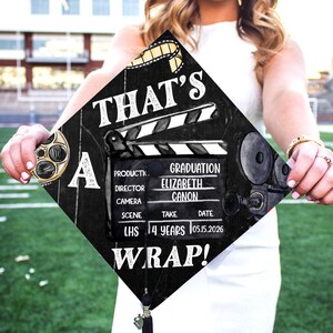 May include: Black graduation cap with a film theme. The cap features a clapperboard design with the text "THAT'S A WRAP!" and graduation details. A film reel and a vintage camera are also depicted.