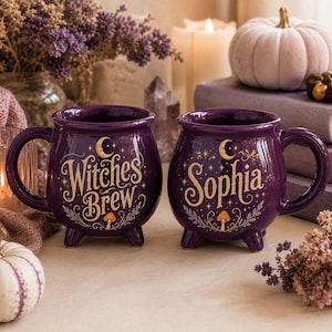 Könnte beinhalten: Zwei violette Keramiktassen in Kesselform mit drei kleinen Füßen. Eine Tasse zeigt den goldenen Schriftzug Witches Brew, eine Mondsichel, Sterne und einen Pilz. Die andere Tasse zeigt den goldenen Namen Sophia, eine Mondsichel und einen Pilz.