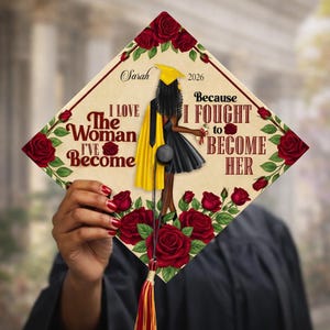 May include: A graduation cap decorated with a design of a woman in a black dress and yellow cape, framed by red roses. The cap displays the text "I LOVE The Woman I'VE Become" and "Because I FOUGHT to BECOME HER".