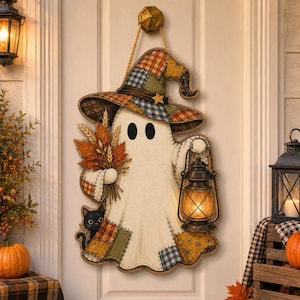 May include: A hanging wall decoration shaped like a ghost wearing a patchwork witch hat. The ghost holds a bundle of autumn leaves and a lit lantern, with a small black cat peeking from the bottom. The design features orange, brown, and blue tones.