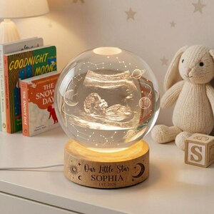 May include: A spherical glass night light featuring an engraved ultrasound image, planets, and constellations. The light sits on a circular wooden base engraved with the text "Our Little Star SOPHIA EST. 2026" along with sun and moon icons.