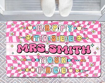 May include: A rectangular doormat with a pink and white checkered design. The mat displays the text "HAPPY TO SEE MRS. SMITH YOUR FACE" in colourful bubble letters, with decorative flowers, apples, and smiley faces. The mat is designed for a teacher.