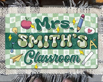 May include: A rectangular doormat with a green and white chequered pattern. The mat features the text "Mrs. Smith's Classroom" in a retro font, along with illustrations of an apple, pencil, and other school-related icons. The mat is placed on a patterned rug.