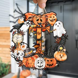 May include: A Halloween-themed wreath with an orange bow, pumpkins, ghosts, and a letter "T". The wreath features a variety of spooky elements, including a skull, a clock, and a crow. The design is colorful and festive, perfect for seasonal decor.