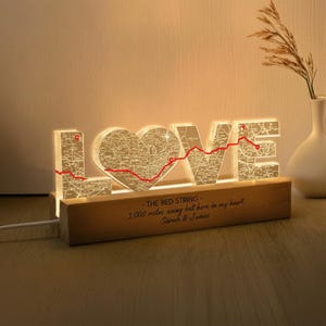 May include: Illuminated "LOVE" sign with a heart-shaped "O" and map detailing. A red line connects the letters, and the base reads "THE RED STRING - 3,000 miles away but here in my heart. Sarah & James."
