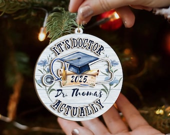 May include: A round wooden Christmas ornament with the text "IT'S DOCTOR ACTUALLY" and "2025 Dr. Thomas". The ornament features a blue graduation cap, a stethoscope, and floral accents. The ornament is being held by a person.