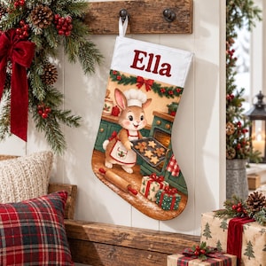 May include: A white Christmas stocking featuring the name Ella in red text at the top. The design depicts a cartoon rabbit wearing a chef hat and apron, pulling a tray of star-shaped cookies from an oven in a kitchen setting with gifts.