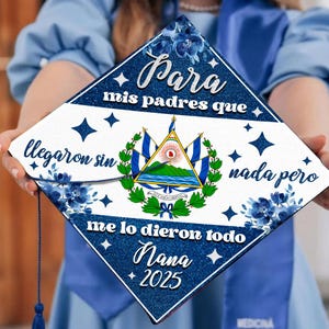 May include: A blue and white graduation cap with a white background and blue trim. The cap features the El Salvadorian flag and the text "Para mis padres que llegaron sin nada pero me lo dieron todo Nana 2025".