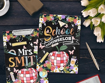 May include: Two clipboards with floral designs and the text "School Counselor" and "Mrs. SMITH". Each clipboard features a red and pink apple design, butterflies, and school-themed illustrations. The clipboards are on a dark wooden surface.