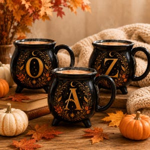 May include: Three black ceramic mugs shaped like cauldrons, each featuring a gold letter O, Z, or A surrounded by an intricate botanical wreath design with stars and crescent moons. The mugs have three small feet and a single handle.