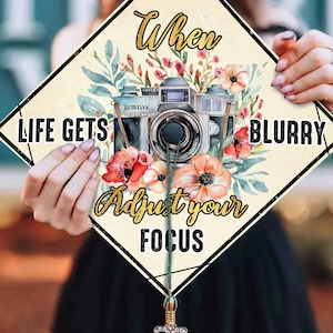 May include: A graduation cap design with the text "When Life Gets Blurry Adjust Your Focus." The design features a vintage camera illustration with floral accents in shades of red, orange, and green.