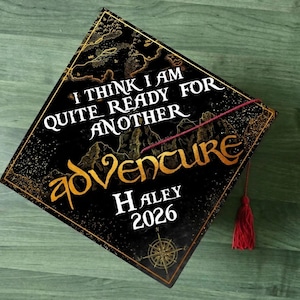 May include: Black graduation cap with gold accents and the words "I THINK I AM QUITE READY FOR ANOTHER ADVENTURE" in white and gold. Includes the name "Haley" and the year "2026". A red tassel hangs from the cap.