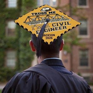 May include: A graduation cap with a yellow and black design. The cap reads "TRUSS ME" and "I'M A CIVIL ENGINEER JACKSON 2026" with a bridge graphic. A black tassel hangs from the cap.