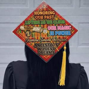 May include: A graduation cap with a colorful design and text. The cap features the phrases "HONORING OUR PAST," "LIFTING AS WE CLIMB," and "OUR MAGIC IS POWER." It also includes the name "Jasmine Simone Thompson" and "class of 2026."