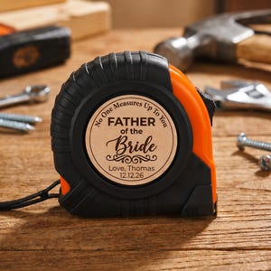 May include: Black and orange tape measure with the text "No One Measures Up To You, FATHER of the Bride, Love, Thomas 12.12.26". The tape measure is on a wooden surface with tools in the background.