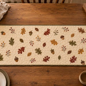 May include: A rectangular quilted table runner with an off-white background featuring a repeating pattern of embroidered autumn leaves, acorns, and berry sprigs in shades of red, orange, yellow, and green, finished with a dark green fabric border.