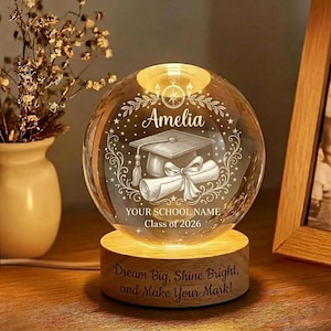 May include: A clear glass graduation sphere with the name "Amelia", a mortarboard, and a diploma etched inside. The sphere sits on a wooden base with the text "Dream Big, Shine Bright, and Make Your Mark!". The base is illuminated with a warm glow.