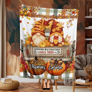Può includere: Una coperta a tema autunnale con gnomi, zucche e foglie autunnali. Il design include un camion con la scritta "Growing Old Together Since 1960". Due zucche sono personalizzate con i nomi "Thomas" e "Emma".