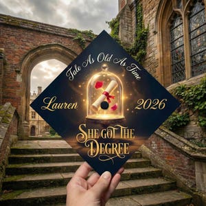 May include: A graduation cap with the text "Tale As Old As Time" and "Lauren 2026". The cap features a Beauty and the Beast theme with a scroll inside a glass dome and the words "She Got The Degree".