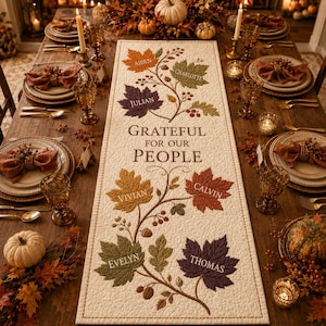 May include: A rectangular cream-colored fabric table runner featuring an embroidered design of autumn leaves in shades of orange, green, purple, and brown. The text reads Grateful for our people, with names Aiden, Charlotte, Julian, Vivian, Calvin, Evelyn, and Thomas.