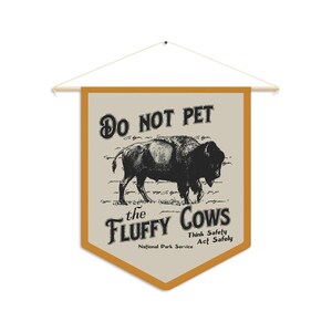 May include: A tan and orange pennant flag with the text "Do Not Pet the Fluffy Cows" and a black and white illustration of a bison. The text "Think Safety Act Safely" and "National Park Service" are also included.