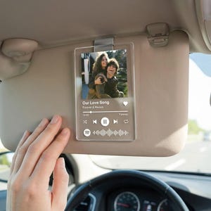 Peut inclure: Un accessoire de pare-soleil de voiture en acrylique transparent avec une photo d'un couple et des détails musicaux. Le texte "Our Love Song" et "Forever & Always" est visible. Le pare-soleil est fixé à un pare-soleil de voiture.