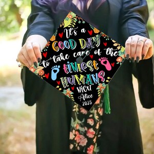 May include: A black graduation cap with colorful floral and heart accents. The cap features the text "It's a GOOD DAY to take care of the tiniest HUMANS" and "NICU Alice 2025". A green tassel hangs from the cap.