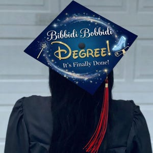 Op de afbeelding: Een marineblauwe afstudeerhoed met gouden tekst "Bibbidi Bobbidi Degree! It's Finally Done!". De hoed heeft een toverstaf, een glazen muiltje en een rol. Een rode kwast hangt aan de hoed.