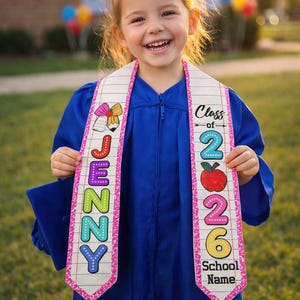 May include: A young child in a blue graduation gown smiles, wearing a white sash with the name "JENNY" in colorful letters. The sash also features the text "Class of 2026" and a red apple graphic, celebrating a school milestone.