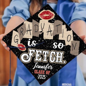 Puede incluir: Gorra de graduación negra con el texto "GRADUATION is so FETCH." y el nombre "Jennifer" y "CLASS OF 2025." La gorra tiene un fondo estrellado y gráficos de labios rojos.