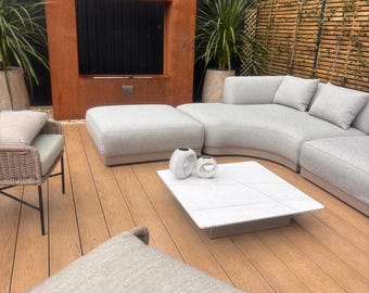 May include: A curved light gray outdoor sectional sofa with a matching ottoman, a low square white coffee table with a grid pattern, and two white ceramic decorative objects. The furniture is arranged on a wooden deck with a large rust-colored frame.