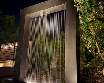 May include: A rectangular dark metal water feature frame with a vertical curtain of water falling from the top beam into a base basin. The structure is illuminated from below, highlighting the water streams against a dark night background.