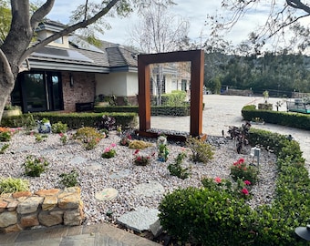 May include: A large rectangular rust-colored metal water feature stands in a garden bed filled with light-colored gravel, stone pavers, and small green shrubs with pink flowers. The structure has a thin stream of water falling from the top frame.