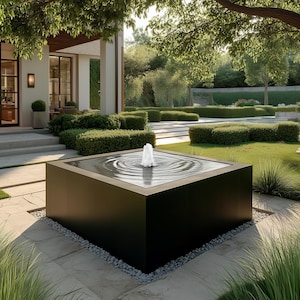 May include: A contemporary outdoor water feature with a dark square basin. A water jet rises from the centre, creating ripples. The fountain is set in a landscaped garden with green grass, shrubs, and a large tree.