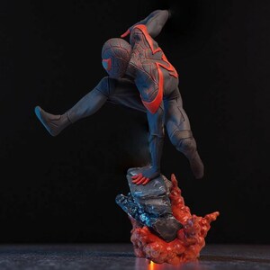 May include: A dynamic figurine of a superhero in a black and red suit, poised mid-action. The figure is atop a rocky base with fiery red accents, set against a dark background. The superhero is in a crouching position.