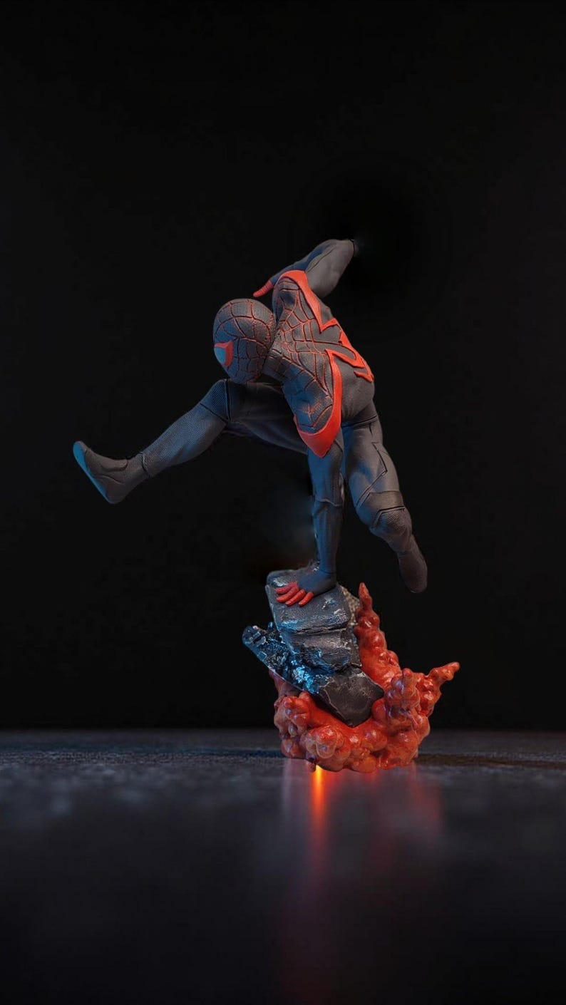 May include: A dynamic figurine of a superhero in a black and red suit, poised mid-action. The figure is atop a rocky base with fiery red accents, set against a dark background. The superhero is in a crouching position.