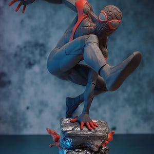 May include: A dynamic figurine of a superhero in a crouching pose, clad in a gray and black suit with red accents. The figure is perched atop a textured, gray base with red, flame-like details, set against a blue-toned backdrop.