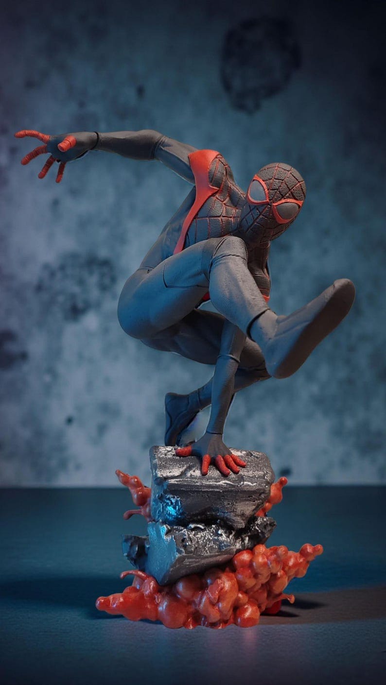 May include: A dynamic figurine of a superhero in a crouching pose, clad in a gray and black suit with red accents. The figure is perched atop a textured, gray base with red, flame-like details, set against a blue-toned backdrop.