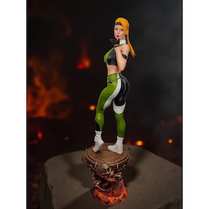May include: A detailed figurine of a female character in a fighting pose. She wears a black and green outfit with white accents, and white boots. Her blonde hair is pulled back. The figurine stands on a decorative base with a skull motif.