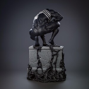 May include: A black and white Venom statue crouching on a gray stone base with black tendrils. The statue is detailed with a textured surface and a white spider symbol on the back.