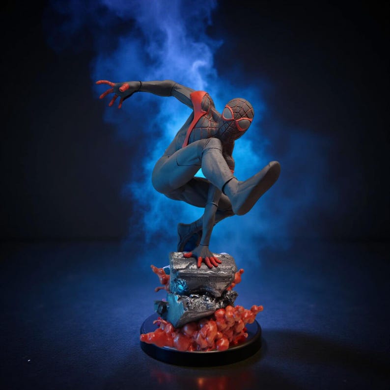May include: A dynamic figurine of a superhero in a leaping pose. The figure is predominantly black with red accents on the hands, mask, and chest. It stands on a detailed base with a dark background and blue smoke effects.