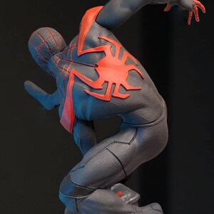 May include: A dynamic statue of a superhero in a black and red suit, poised in a leaping pose. The figure has a detailed spider-web design on the head and a large red spider emblem on the back. The base is a textured, rocky surface.