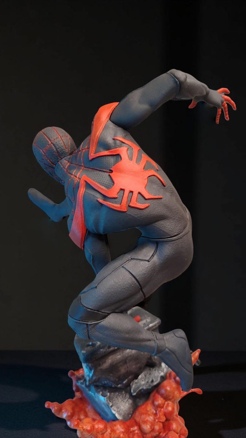 May include: A dynamic statue of a superhero in a black and red suit, poised in a leaping pose. The figure has a detailed spider-web design on the head and a large red spider emblem on the back. The base is a textured, rocky surface.