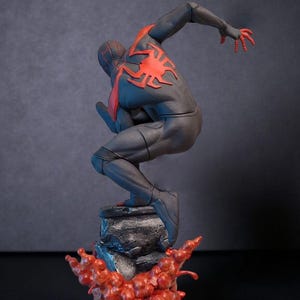 May include: A black and red figurine of a superhero in a dynamic pose, perched atop a rocky base with a red, flame-like effect. The figure has a red spider emblem on the back.