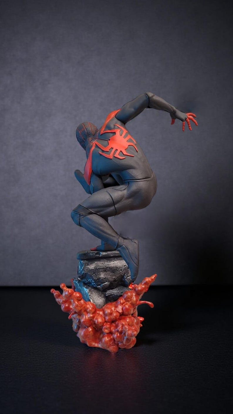 May include: A black and red figurine of a superhero in a dynamic pose, perched atop a rocky base with a red, flame-like effect. The figure has a red spider emblem on the back.